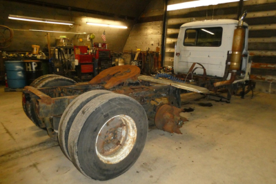 Truck chassis and cab in a workshop; derelict 1985 Mack R600 truck with exposed frame, large dual rear wheels, and a white cab in background.