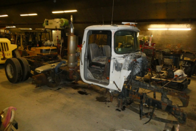 Mack R600 truck cab in workshop showing damaged front end and exposed chassis.