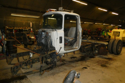 Mack R600 truck cab and chassis in a workshop showing exposed frame and missing bed, with scattered parts on the floor.