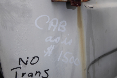 Scribbled text and markings on a vehicle surface. Visible words include 'CAB', 'NO Trans', '1500', and some handwritten notes in white/gray chalk on a light gray metal panel; rust line visible. 