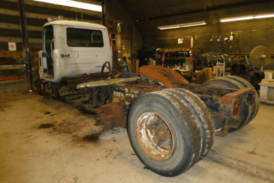 Rear view of a Mack R600 conventional daycab chassis in a workshop, showing dual rear wheels, chassis frame, and cab in background.