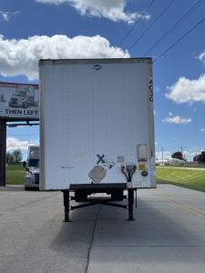 Rear view of a white utility trailer with support legs down, showing no visible branding and a couple of markings on the lower left corner. Clear blue sky with clouds.