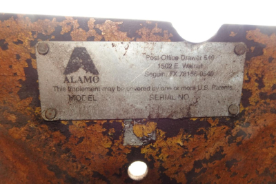 Metal nameplate on equipment showing Alamo logo and address, with serial number and patent text. Weathered metal with rust patina and screws at corners.