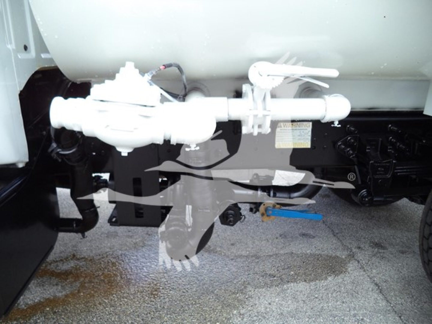 Truck undercarriage showing fuel line assembly and valve near cylindrical tank, with label and blue tool on ground; white plumbing components attached to black chassis.