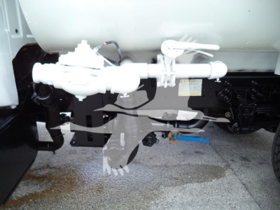 Truck undercarriage showing fuel line assembly and valve near cylindrical tank, with label and blue tool on ground; white plumbing components attached to black chassis.