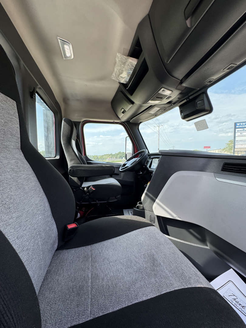 Truck cab interior view of a 2022 Freightliner Cascadia 116 showing driver seat, passenger seat, and dashboard from the passenger side, with sun visors and overhead storage.