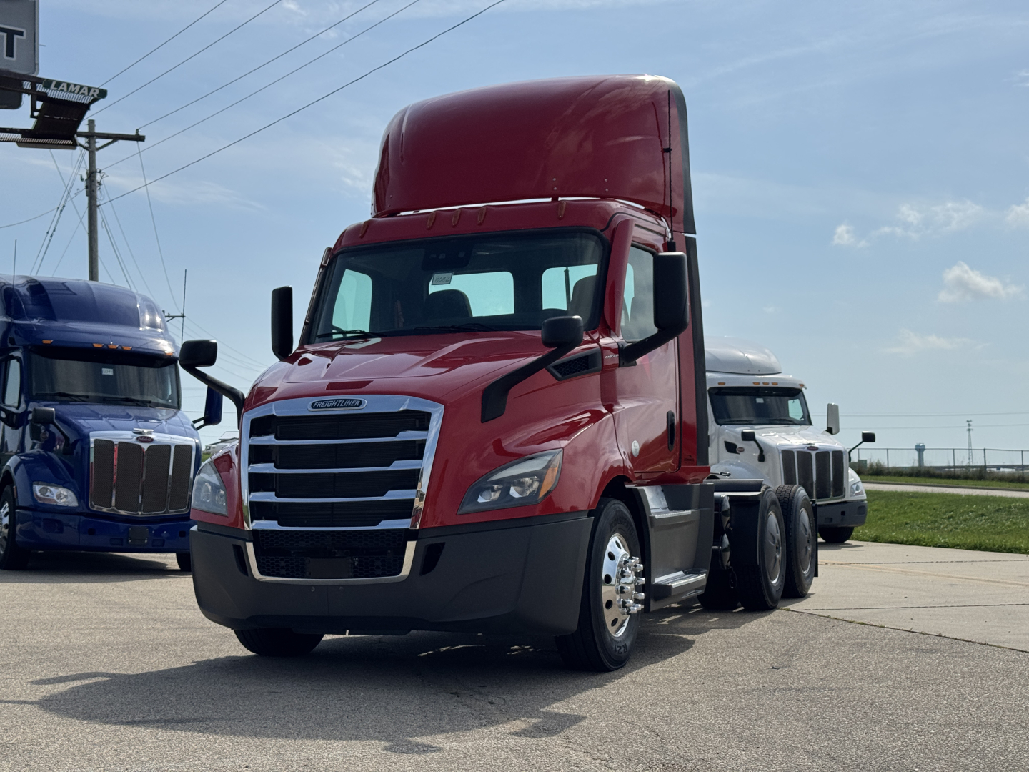 Truck front-left view of a red 2022 Freightliner Cascadia 116 day cab tractor with tall sleeper, chrome grille, black bumper, and attached blue and white trucks in background.