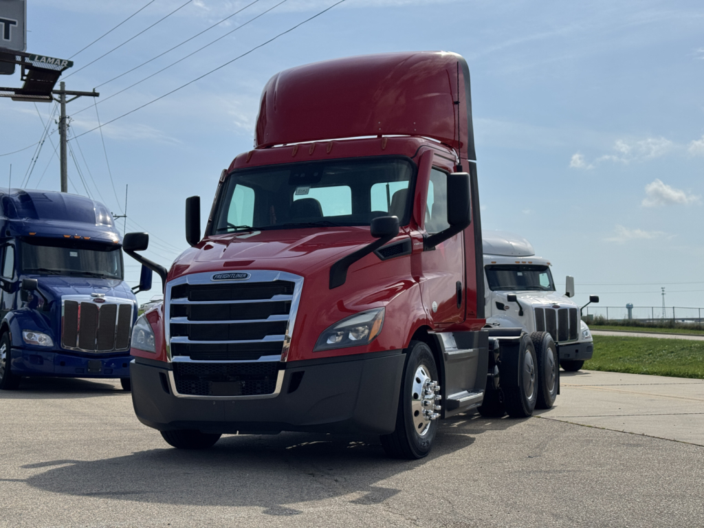 Truck front-left view of a red 2022 Freightliner Cascadia 116 day cab tractor with tall sleeper, chrome grille, black bumper, and attached blue and white trucks in background.