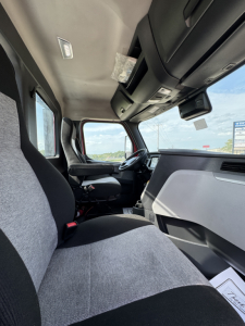 Truck cab interior view of a 2022 Freightliner Cascadia 116 showing driver seat, passenger seat, and dashboard from the passenger side, with sun visors and overhead storage.
