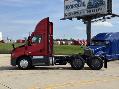 Truck cab viewed from side with red Freightliner Cascadia 116 tractor, black chassis, blue truck in background, billboard sign above.