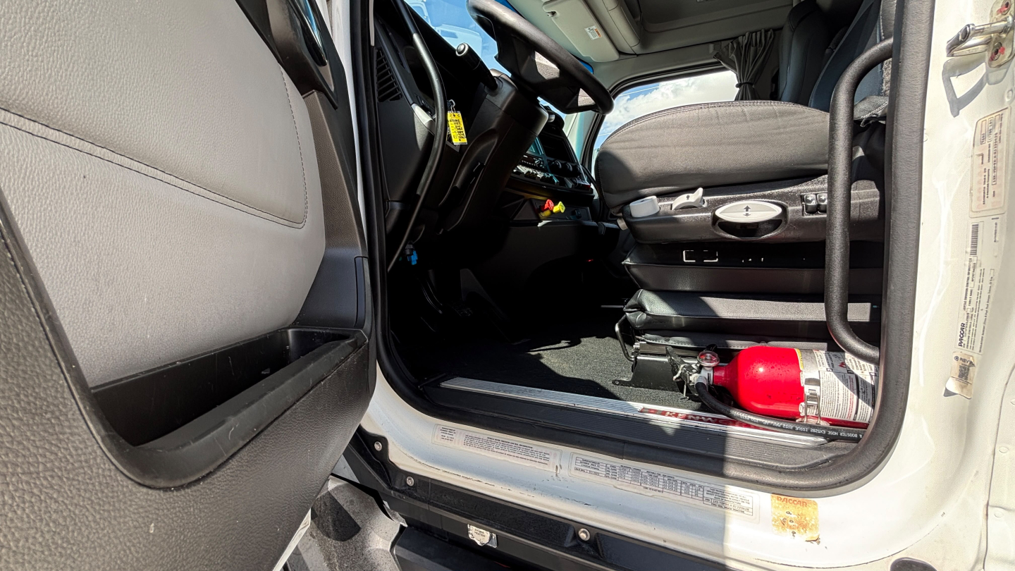 Kenworth T680 cab interior showing open driver door, steering wheel, dashboard, and a red fire extinguisher mounted near the door sill.