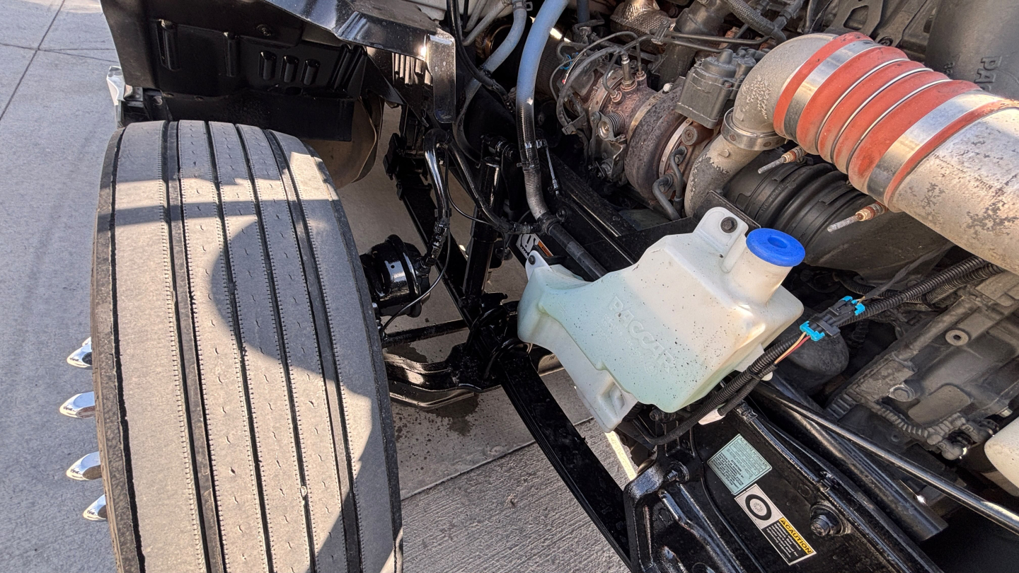 Kenworth T680 engine bay and front wheel area showing coolant reservoir and air intake tube.