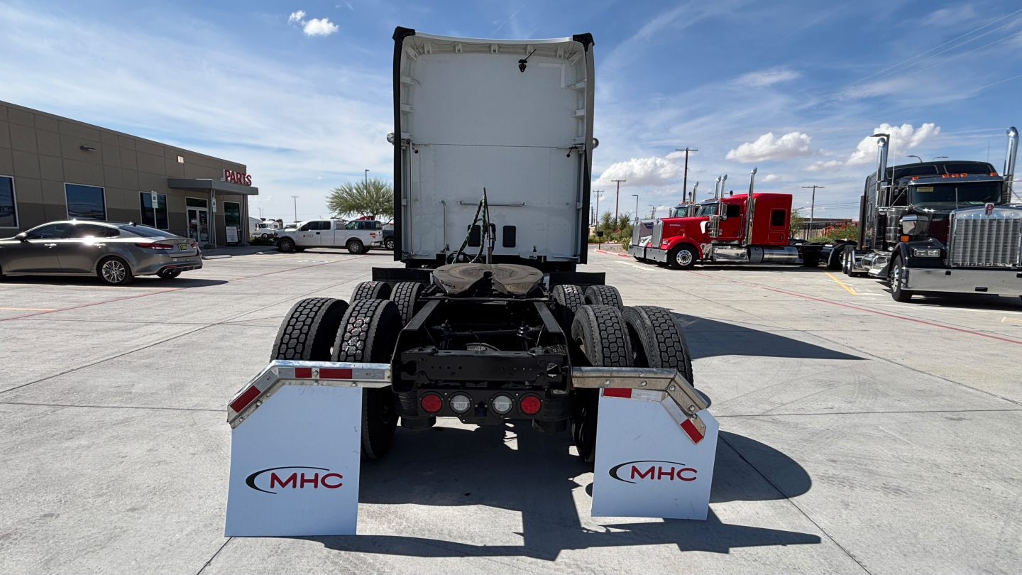 Rear view of a Kenworth T680 semi-truck tractor chassis with mudflaps showing MHC branding, fifth wheel and rear tires visible.