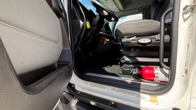 Kenworth T680 cab interior showing open driver door, steering wheel, dashboard, and a red fire extinguisher mounted near the door sill.