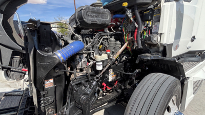 Kenworth T680 engine bay view with exposed components, hoses, and wiring; visible air intake hose, battery area, and radiator support in a truck bay