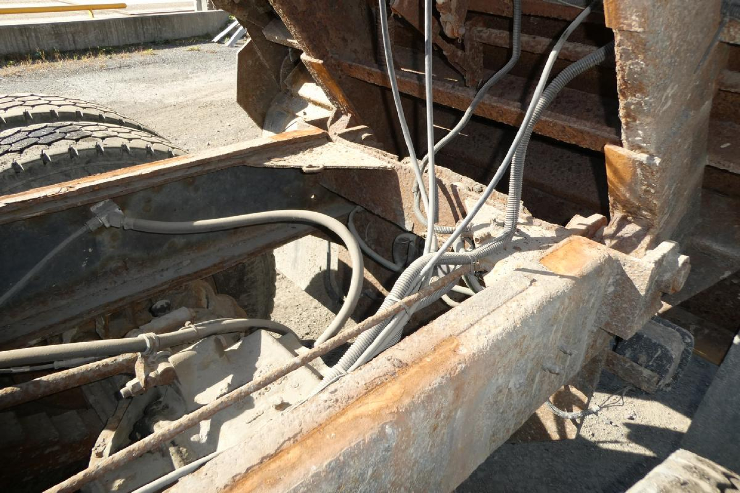 Truck chassis with exposed wiring and rusted frame near rear, hydraulic hoses and electrical cables visible under a dump truck bed.
