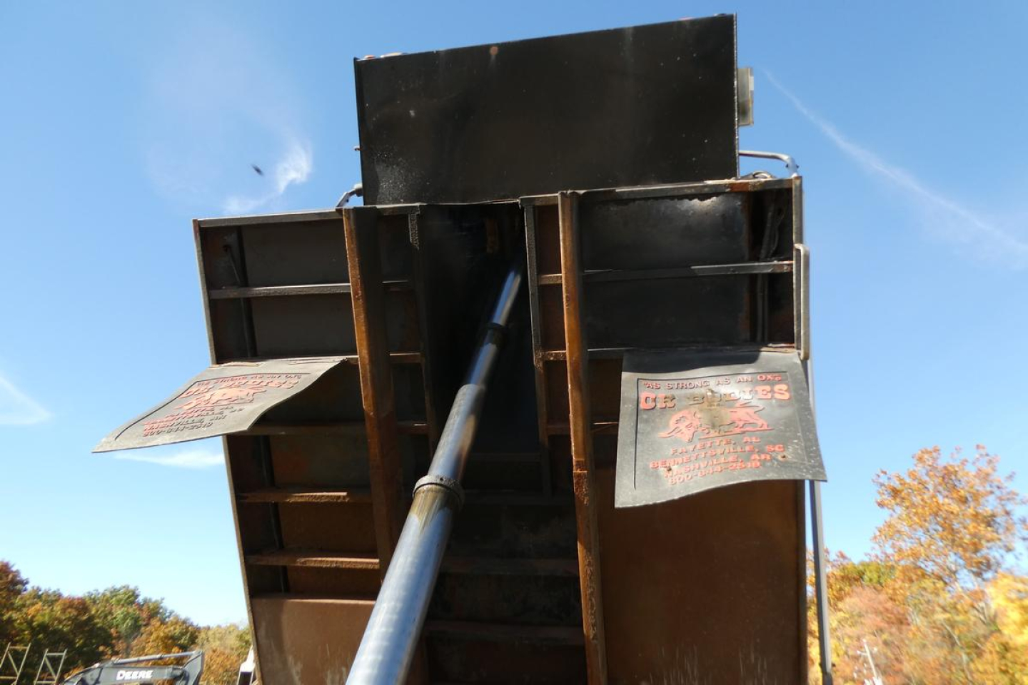 Dump truck bed with hydraulic tailgate and inspection tags visible inside the metal container. Rain of blue sky above, fall foliage in background.