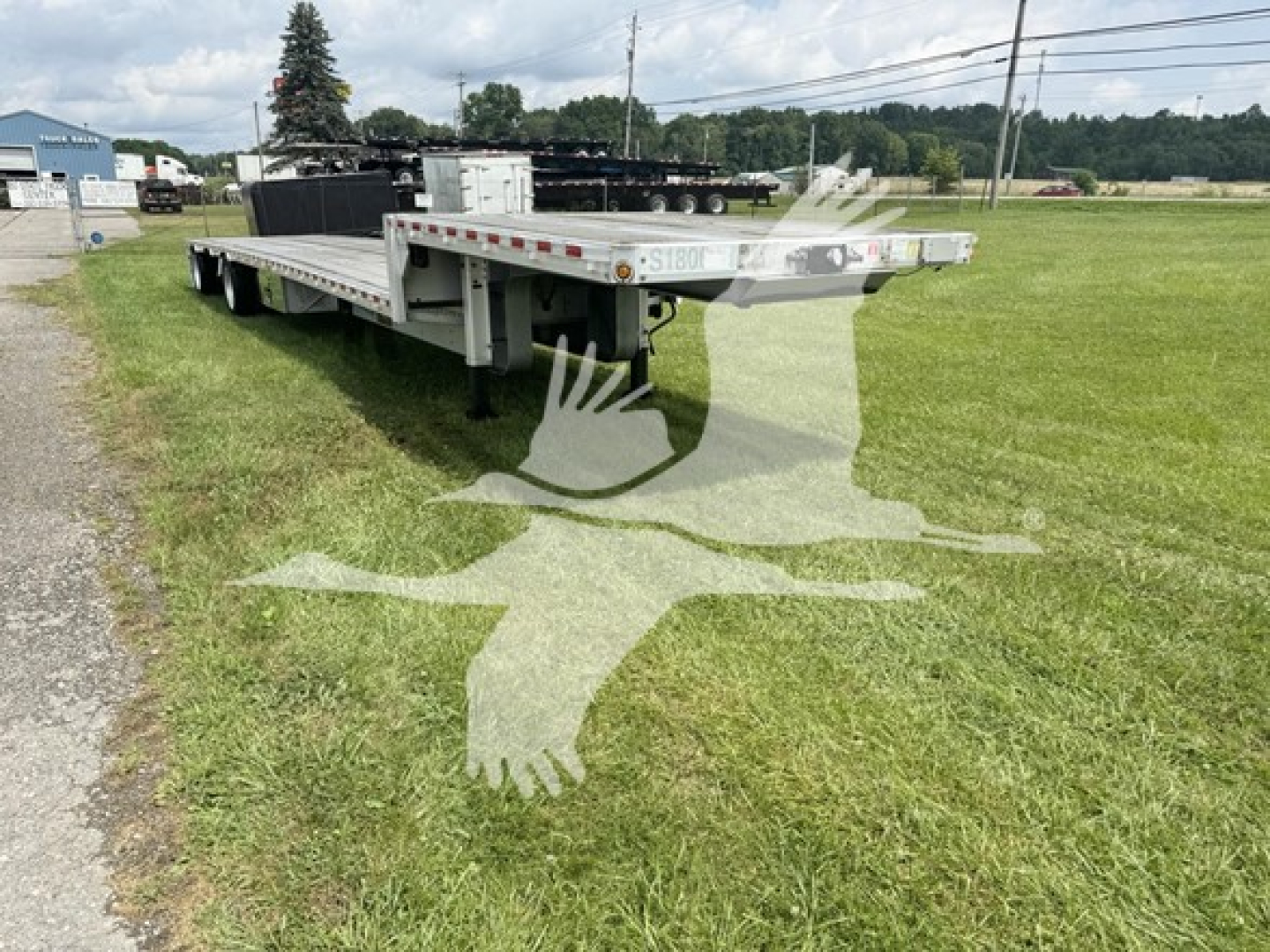 Flatbed trailer with drop deck showing rear and side rails, VIN plate and license plate area, green grass field in background.
