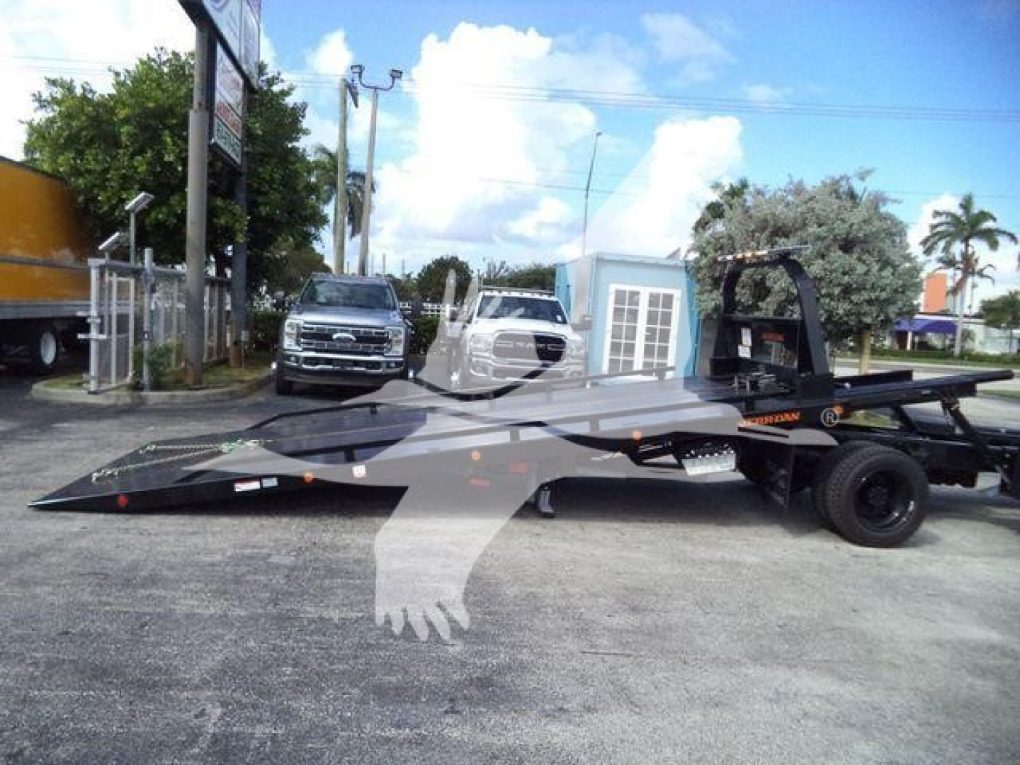 Tow truck rollback with a low-bed trailer and two white trucks in background; Chevrolet Silverado 6500HD listing context visible.