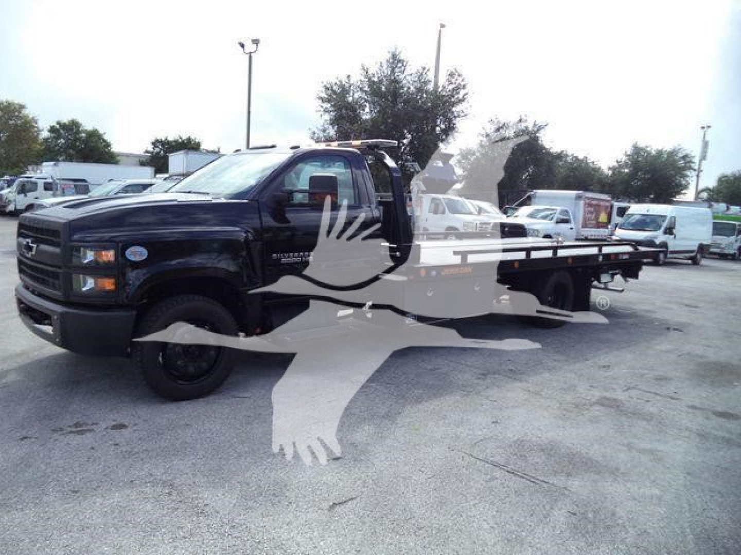 Chevrolet Silverado 6500HD rollback truck in a dealership lot, black cab with flatbed; watermark overlay obscures center.
