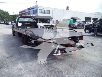 Flatbed tow truck chassis with hydraulic rollback bed, rear view showing metal deck, taillights, and tow hitch on a black trailer in a dealership lot.