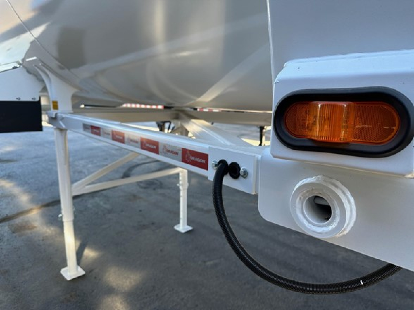 Front left side of a new dump trailer showing a round white access port with a black flexible hose connected, orange side marker light, and white frame with red reflective labels along the side.