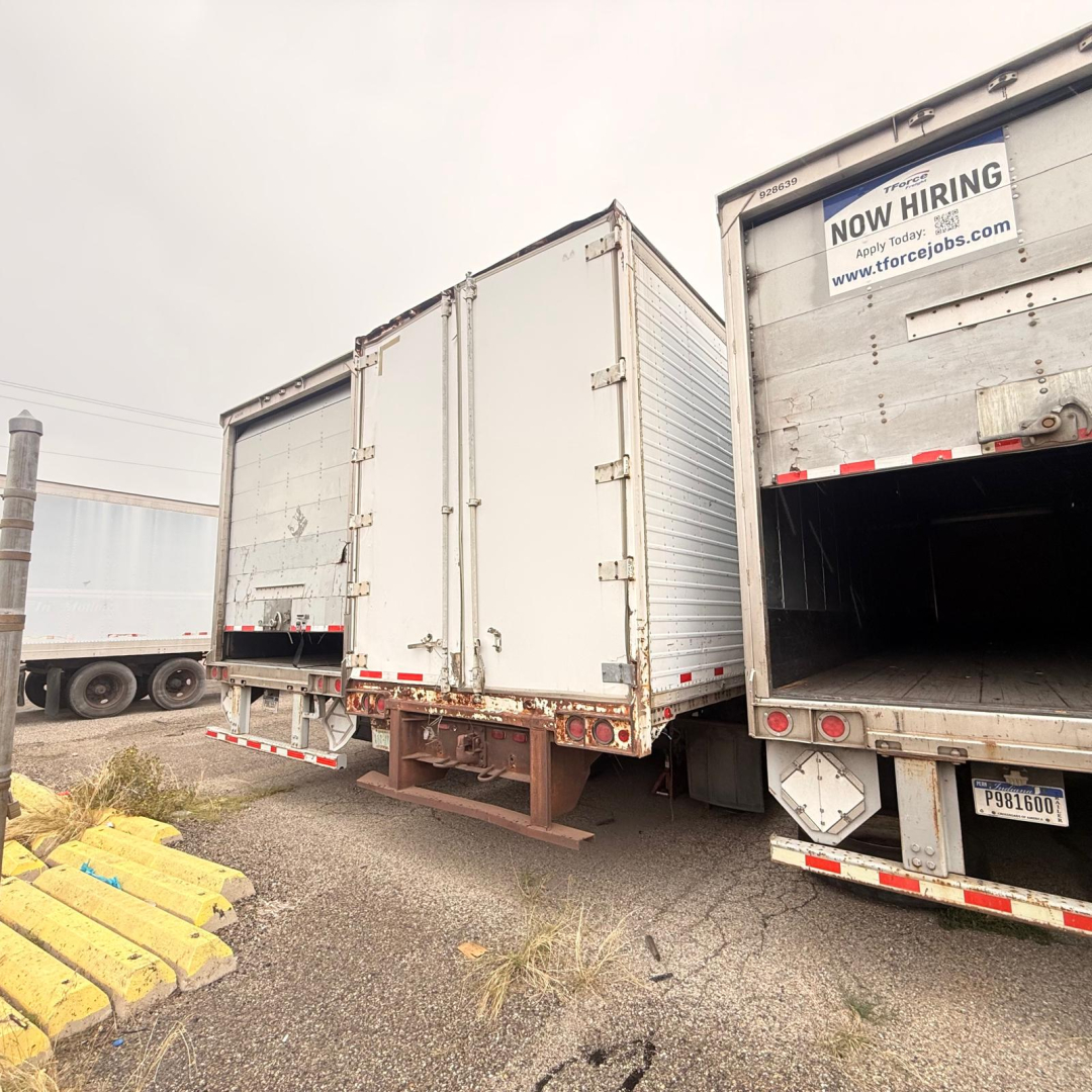Rear view of a white dry van trailer with doors closed, next to an open trailer in a lot. Visible are metal hinges, latches, and bumper with reflective strips; a Now Hiring sign on the adjacent trailer.