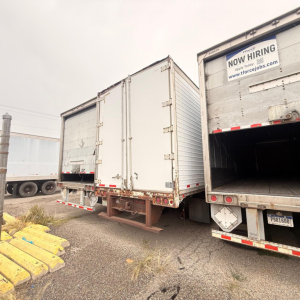 Rear view of a white dry van trailer with doors closed, next to an open trailer in a lot. Visible are metal hinges, latches, and bumper with reflective strips; a Now Hiring sign on the adjacent trailer.