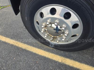 Wheel hub and tire of Freightliner M2 showing polished aluminum hub with multiple lug nuts and reflection of person in pavement.