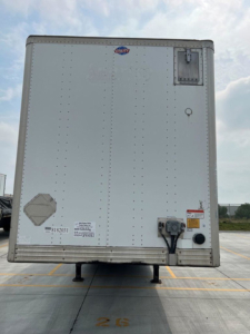 Rear view of a white utility van trailer with a raised floor, black lower landing gear, and wiring connections at the bottom center; a small metal access door on the upper right and a Diamond plate patch on the left rear door.