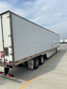 Trailer side view of a long white utility van trailer with dual rear axles and reflective striping along bottom edge.