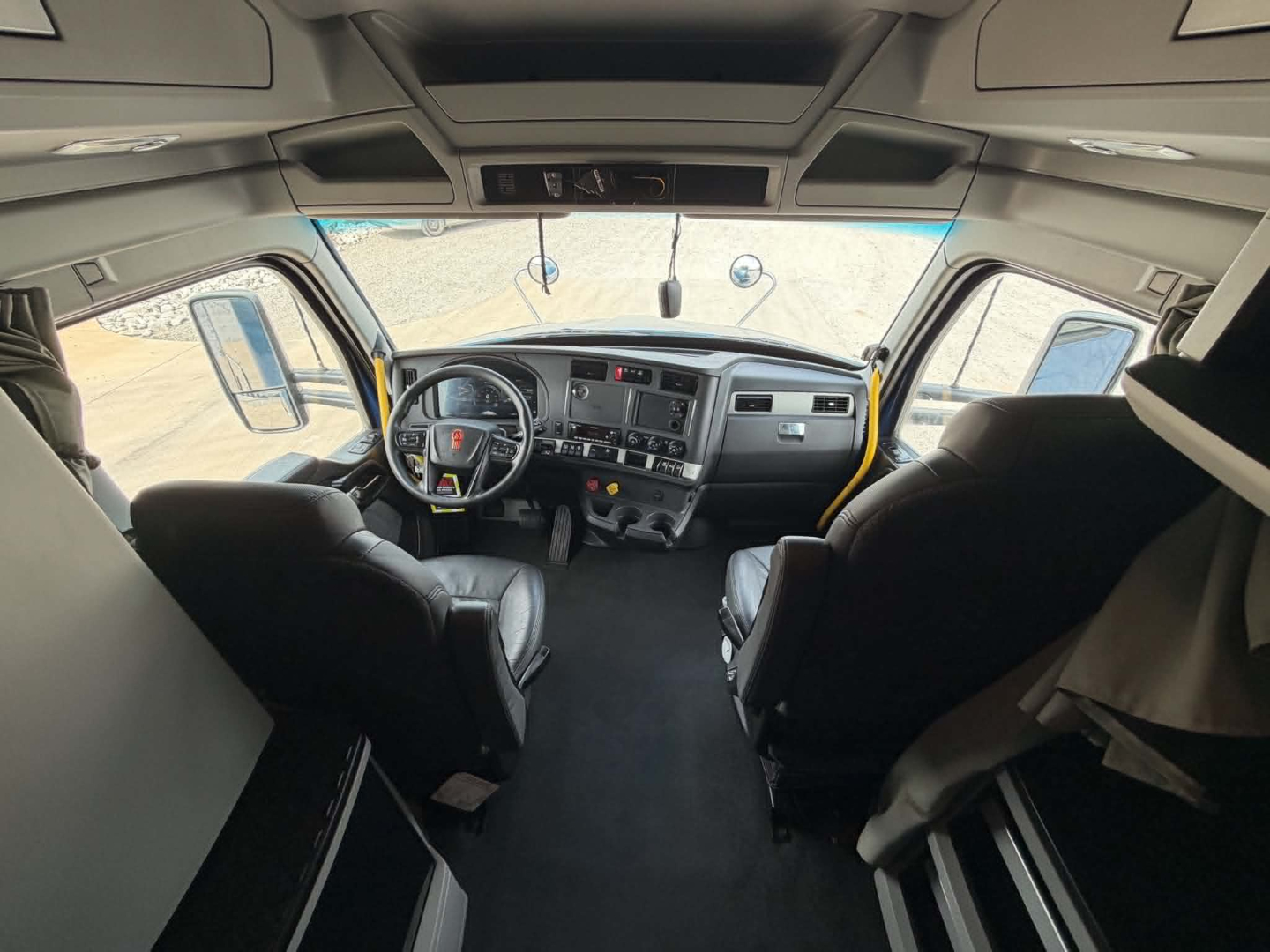Kenworth T680 cockpit view with driver and passenger seats, large dashboard, steering wheel, and center console visible; overhead sun visors and windshield.