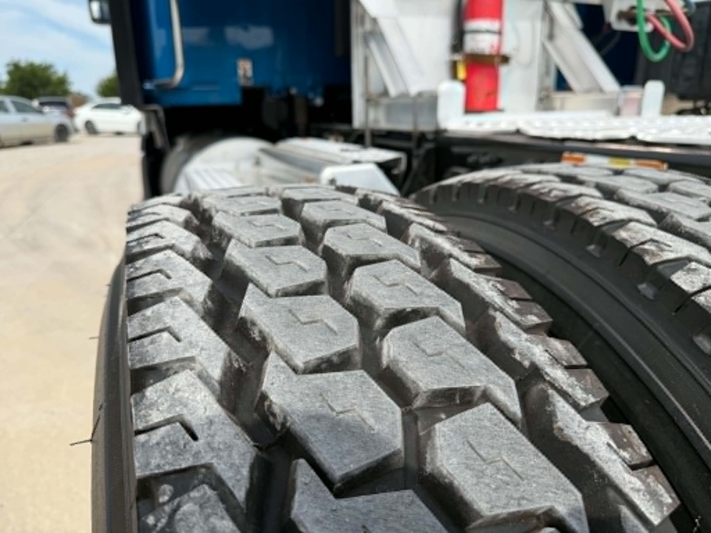 Kenworth truck wheel and dual tires close-up view showing rugged tread pattern and wheel well; part of semi-truck chassis is visible in background.