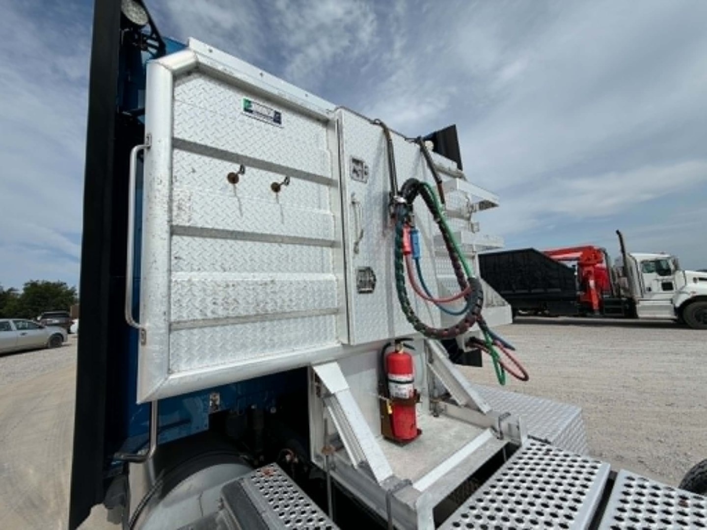 Kenworth T680 dump bed tail area showing aluminum toolbox, hydraulic hoses, fire extinguisher, and step pad on a trailer truck.
