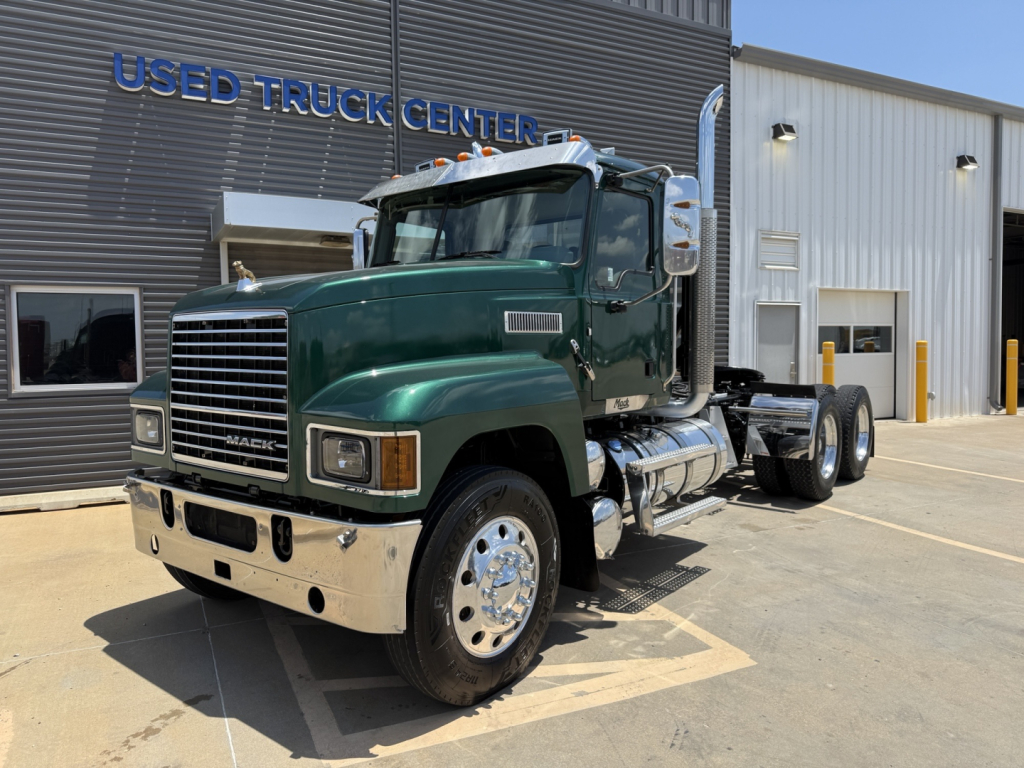 Mack PI64T conventional daycab with green cab and chrome front, dual rear axles, fuel tanks, front grille, and Used Truck Center signage in background.