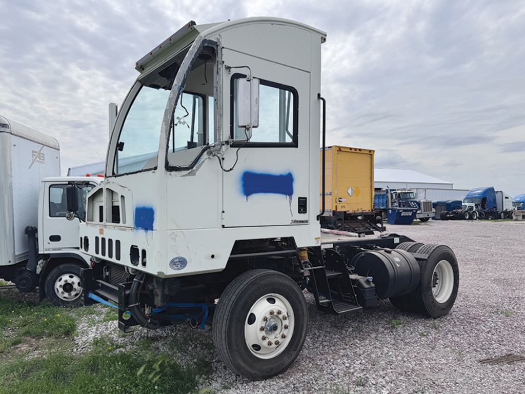 Autocar XSpotter raised roof truck cab chassis in yard, front and side view, white cab with blue patches, dual rear wheels, diesel fuel tank, exposed engine bay, various equipment in background.