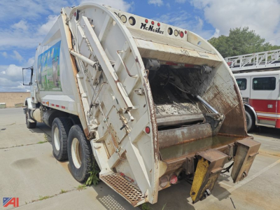 Garbage truck rear view showing compactor door and hydraulic arms, McNeilus branding, three-axle wheel configuration, dirty rear hopper and tipping fork.