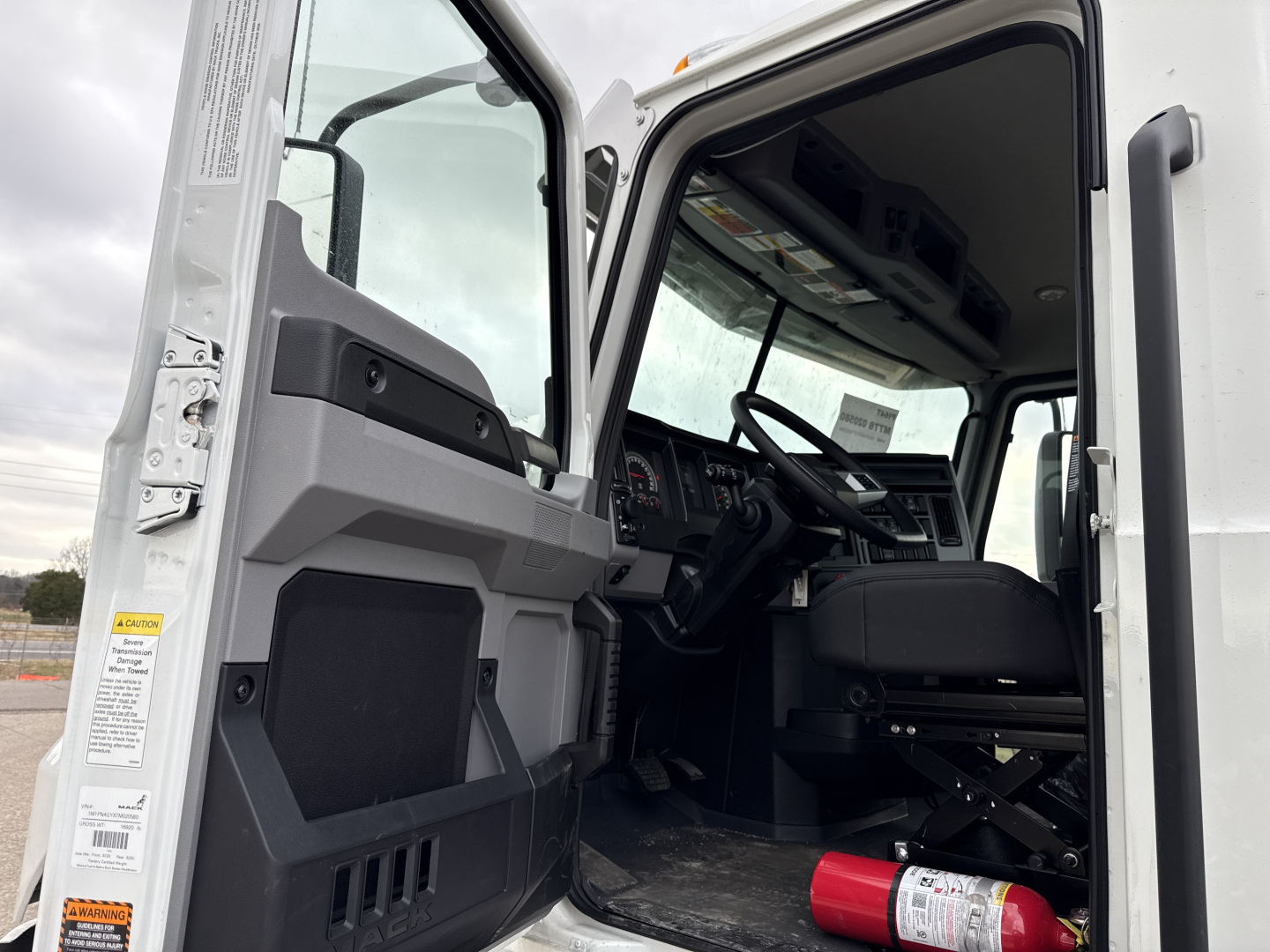 Truck cab interior of a 2026 Mack PI64T showing driver seat, dashboard, steering wheel, and open door with warning labels visible.