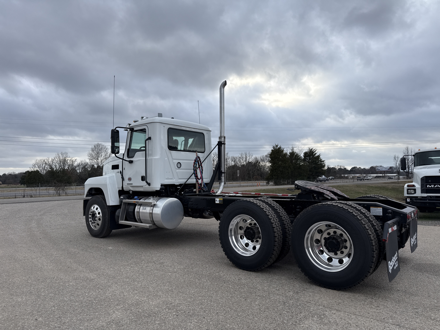 Truck cab and rear chassis of a 2026 Mack PI64T conventional daycab shown from left rear angle; white cab, dual rear axles, exposed fuelTank, exhaust stack, and chassis with fifth wheel.