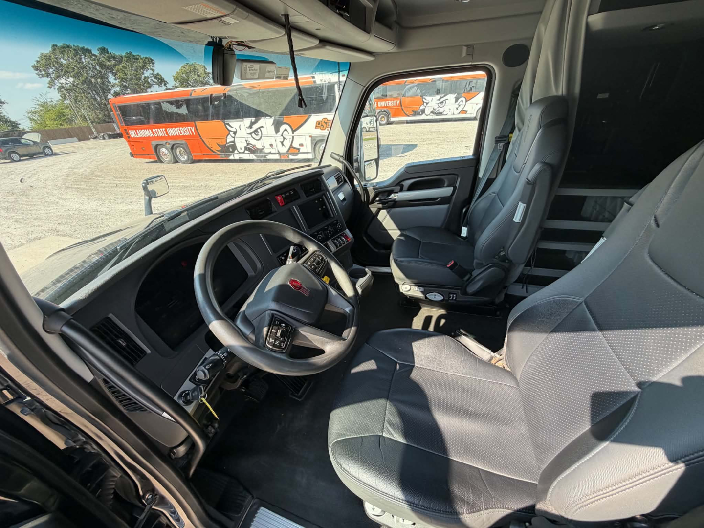Kenworth T680 truck cab interior showing steering wheel, driver seat, and dashboard with digital displays. Exterior lot with orange trucks visible through windshield.