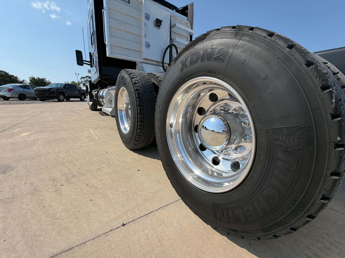 Kenworth T680 wheel and front tire view with chrome hubcap, side steps, and cab behind on a sunny lot.