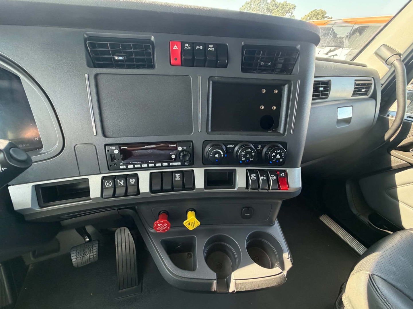 Kenworth T680 cab interior dashboard view showing steering column, center console with climate controls, two cup holders, gear shifter, and red hazard button.