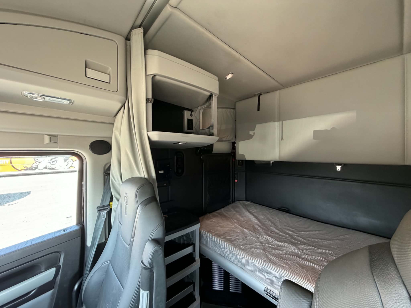 Cab interior of a Kenworth T680 sleeper showing upper bunk, storage cabinets, and driver's seat with a folded bed behind; light gray interior panels.