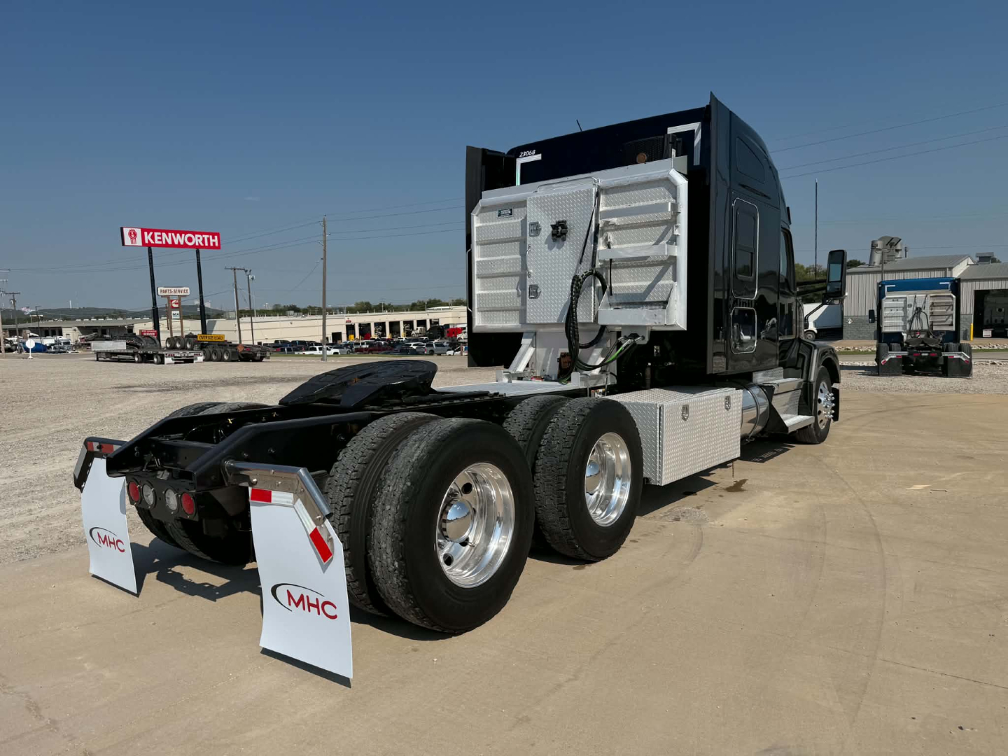 Kenworth T680 tractor in a truck lot, rear view showing aluminum deck, fifth wheel, dual rear axles, chrome wheels, cab with sleeper, rear mudflaps with MHC logos.