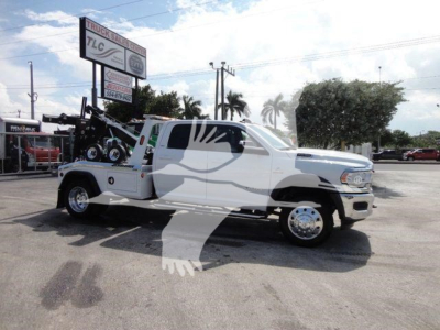 Ram 5500 tow truck with a white cab and extended bed carrying a winch, side view showing front chrome wheels and heavy equipment mounted at rear.