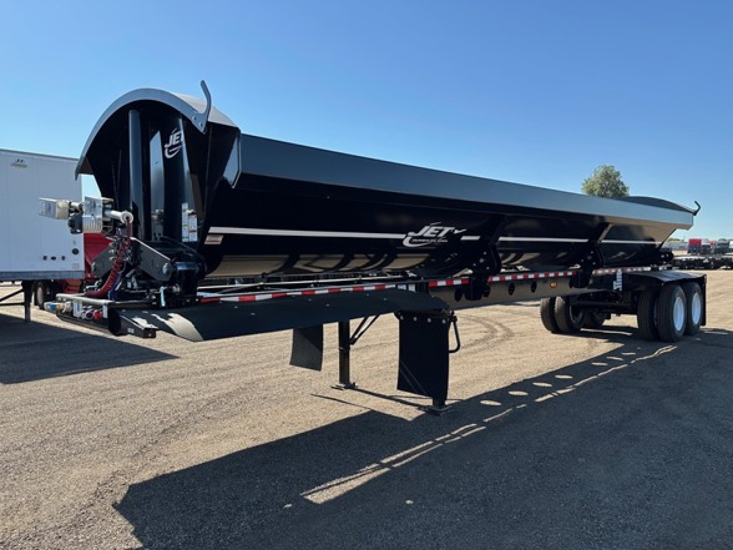 Jet Jet side dump trailer in black, side view showing long rectangular dump body on a lowboy trailer with landing gear, wheels, and hydraulic hoist.