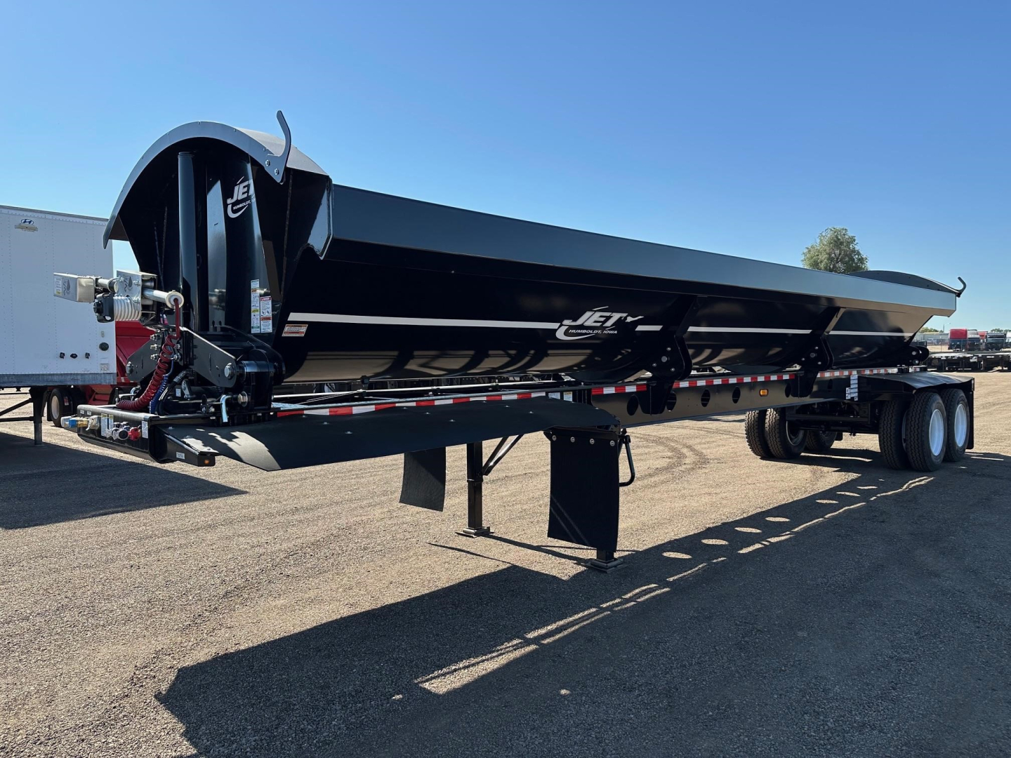 Jet Jet Side Dump trailer showing black steel cargo bed with side discharge, rear dual axle, landing legs, and control mechanism at front left. Visible Jet branding on side.