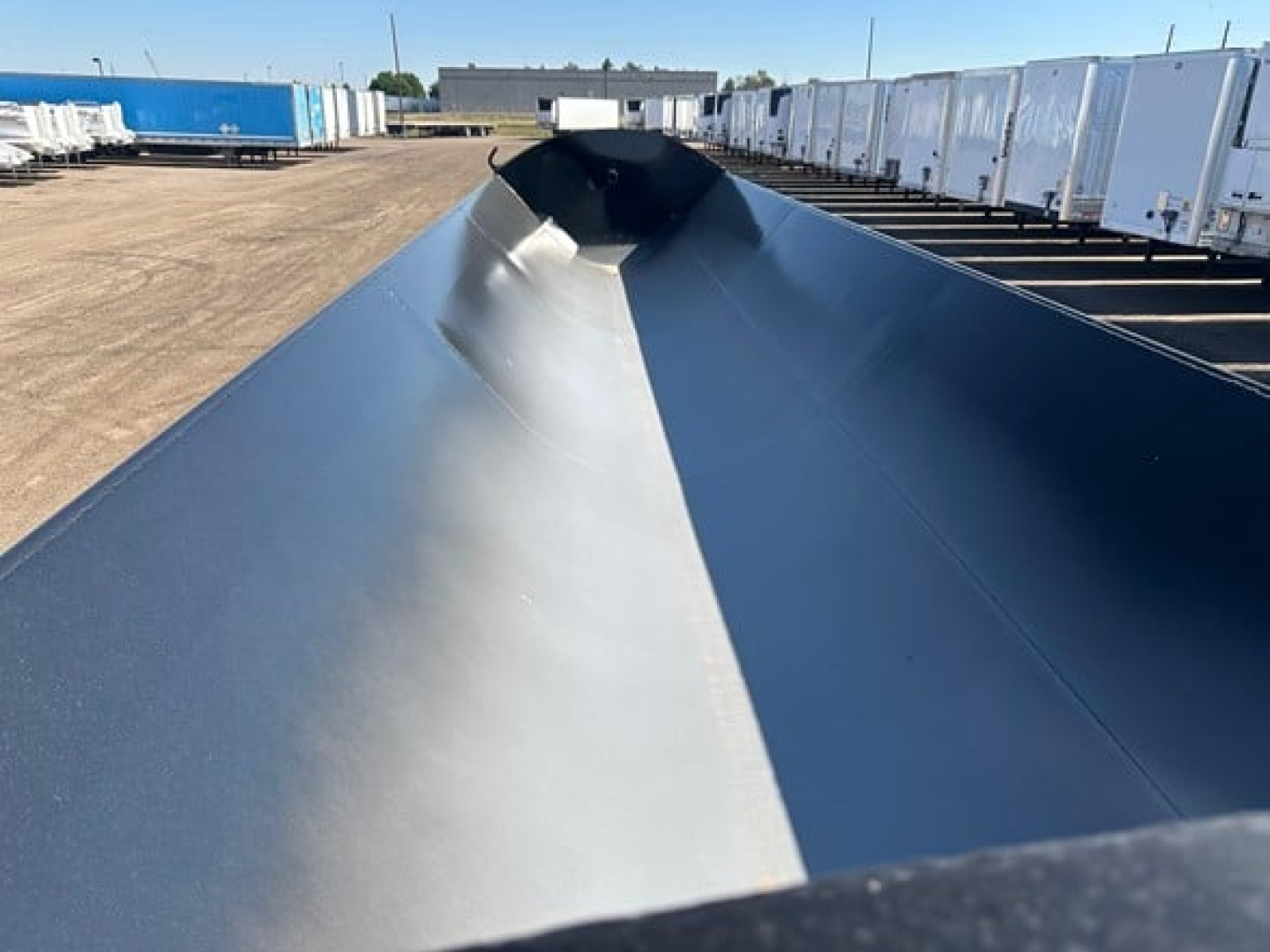 Long metal side dump trailer with high sidewalls, black paint, shot from the top showing a central ridge and reflections, row of trailers in background.