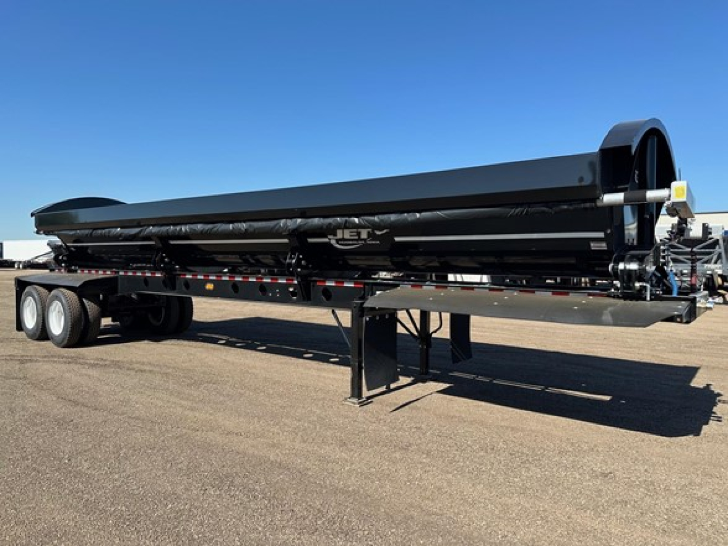 Jet side dump trailer shown in profile with black exterior, tandem axles, and support legs under a long flatbed. Visible branding 'JET' on the side near the center, with storage context around.