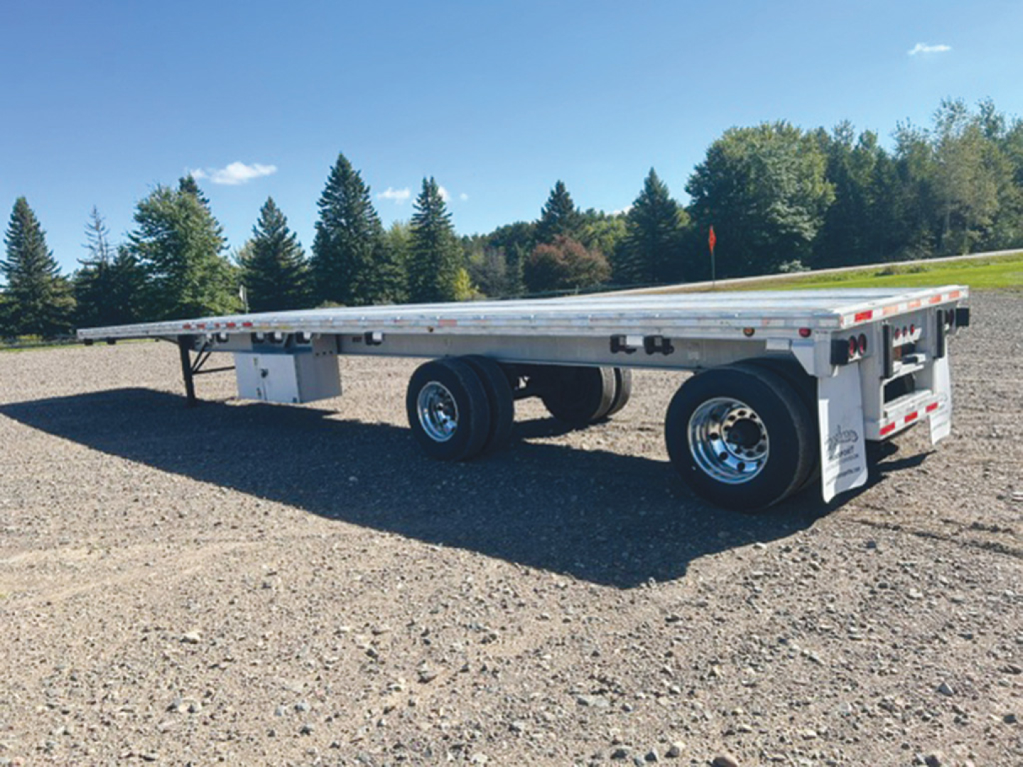 Trailer flatbed showing aluminum deck and dual rear axles with chrome wheels; side rails and rear bumper visible in a gravel lot.